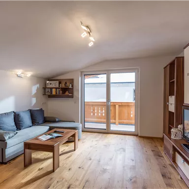 Ein gemütliches Wohnzimmer mit einem grauen Sofa und einem Holztisch. Große Fenster bieten viel Tageslicht und Zugang zum Balkon.