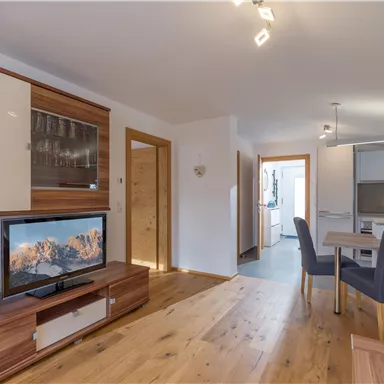 Ein gemütliches Wohnzimmer mit modernem Möbeldesign und einem Fernseher. Der Raum hat helle Farben und einen offenen Zugang zur Küche.