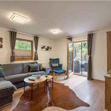 Ein gemütliches Wohnzimmer mit einem Sofa und einem Sessel. Große Fenster bieten einen Blick nach draußen und viel natürliches Licht.