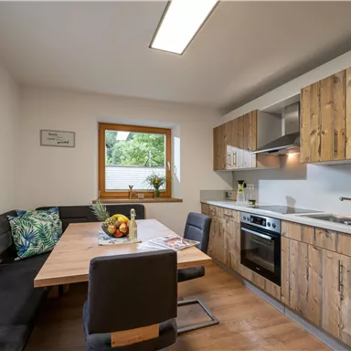 Eine moderne Küche mit Holzmöbeln und einer großen Fensterfront. Am Tisch stehen frisches Obst und eine einladende Sitzbank.