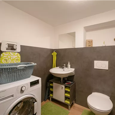 Ein modernes Badezimmer mit einer Waschmaschine, einem Waschbecken und einer hellen Wandgestaltung. Ein Wäschekorb und Pflegeprodukte sind ebenfalls sichtbar.