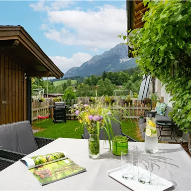 Ein gemütlicher Außenbereich mit einem Tisch, bunten Blumen und Blick auf die Berge. Im Hintergrund sind ein Garten und ein Holzschuppen zu sehen.