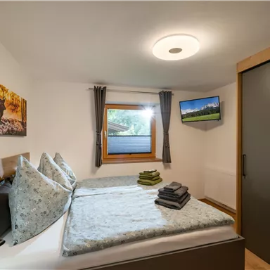 Ein gemütliches Schlafzimmer mit einem Doppelbett, Kissen und sauberen Handtüchern. Ein Fenster bietet Blick nach draußen und ein Fernseher ist an der Wand montiert.