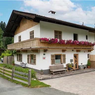 Ein charmantes Haus mit einem Balkon und bunten Blumen. Die Umgebung ist grün und die Straße führt vorbei.