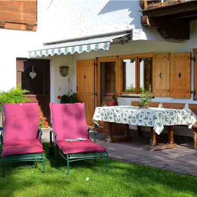 A cozy terrace with two red lounge chairs and a blooming table. In the background, wooden windows and green plants can be seen.
