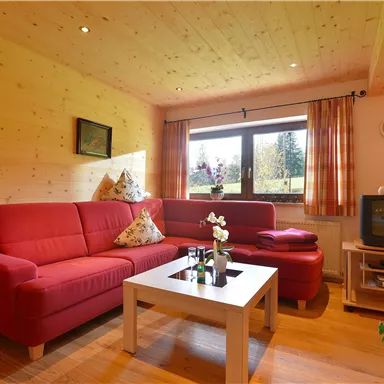 A cozy living room with a red sofa and a white table. Large windows provide plenty of daylight and a view of the greenery.