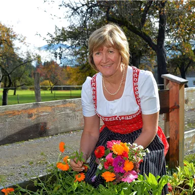 A woman in traditional attire is picking flowers in a garden. In the background, trees and a meadow can be seen.