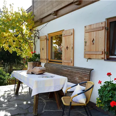 An inviting terrace with a table and chairs in the greenery. The shutters are made of wood and red flowers are blooming.