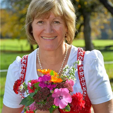 A smiling woman is wearing a traditional dress and holding a colorful bouquet of flowers. In the background, a green meadow landscape and trees can be seen.