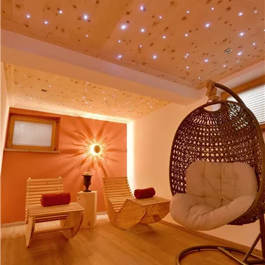 A cozy room with a hanging rocking chair and two wooden armchairs. The walls are painted in warm colors and atmospheric lighting sparkles on the ceiling.