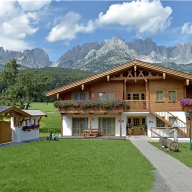 A beautiful wooden house in the middle of a green meadow with colorful flowers. Majestic mountains rise in the background under a blue sky.