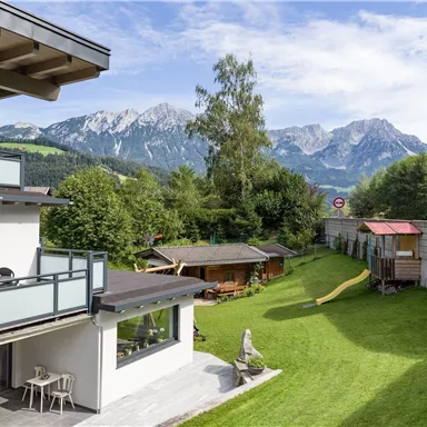 A beautiful garden with a lawn and play areas for children. In the background, impressive mountains and a clear blue sky can be seen.