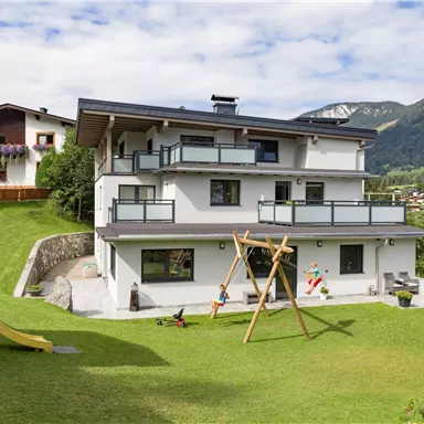 A modern house with several balconies and a large garden. In the garden, there is a swing and a slide, surrounded by mountains and a blue sky.