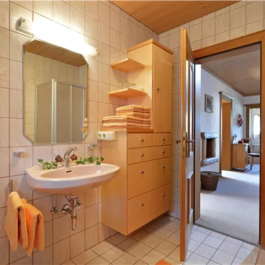 Ein modernes Badezimmer mit einem Waschbecken, Spiegel und einem Holzschrank. Im Hintergrund ist ein Flur mit einem gemütlichen Wohnbereich sichtbar.