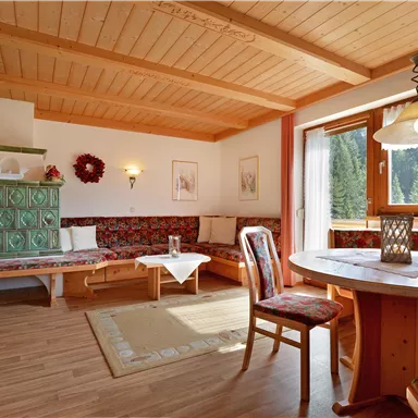 Ein gemütliches Wohnzimmer mit Holzbalkendecke und großen Fenstern. Der Raum ist mit einem Tisch, Stühlen und einer Sitzbank eingerichtet, umgeben von einer ruhigen Naturlandschaft.