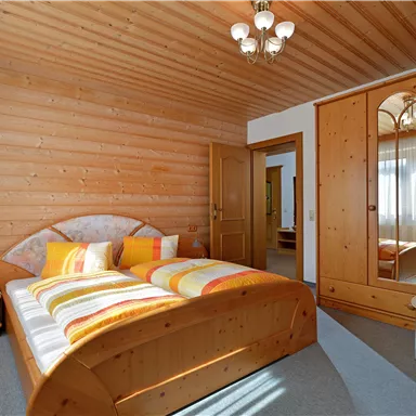 Ein gemütliches Schlafzimmer mit Holzwänden und einem Doppelbett. Die Einrichtung ist hell und freundlich, mit einem großen Kleiderschrank und einem Fenster, das viel Licht hereinlässt.