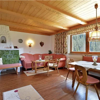 Ein gemütlicher Wohnraum mit Holzdecke und warmen Farben. Die Einrichtung umfasst ein Sofa, einen Tisch und ein großes Fenster mit Blick ins Grüne.