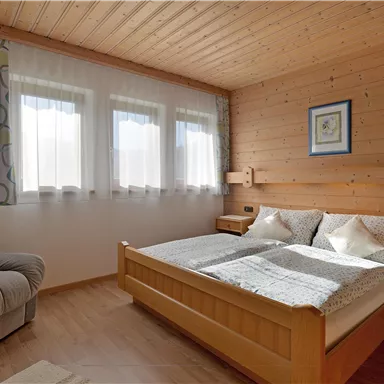 Ein gemütliches Schlafzimmer mit einem Holzbett und einem bequemen Sessel. Große Fenster sorgen für viel Tageslicht.