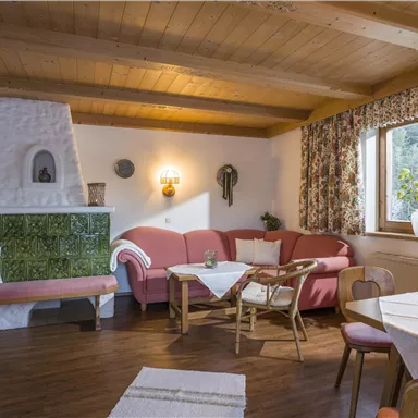 Ein gemütliches Wohnzimmer mit roten Couchgarnituren und einem Holztisch. Große Fenster lassen viel Licht herein und der Holzofen sorgt für eine warme Atmosphäre.