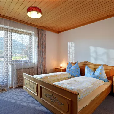 A bright bedroom with a double bed and two blue pillows. Large windows offer a view of the outdoors and let in plenty of light.