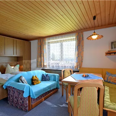 A cozy living space with a double bed, a couch, and a dining area. Large windows provide plenty of daylight.