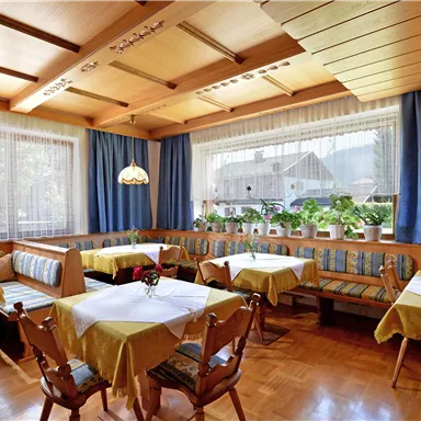 A cozy restaurant with wooden furniture and nicely set tables. There is plenty of light and a green view visible through the windows.