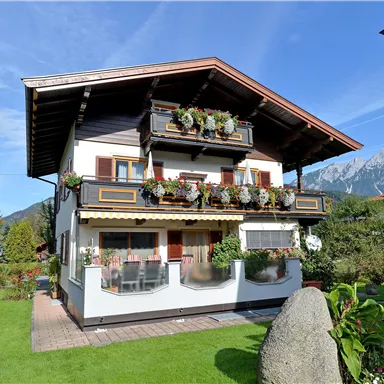 A charming two-story house with flower balconies and a beautiful garden. In the background, mountains and a clear sky are visible.