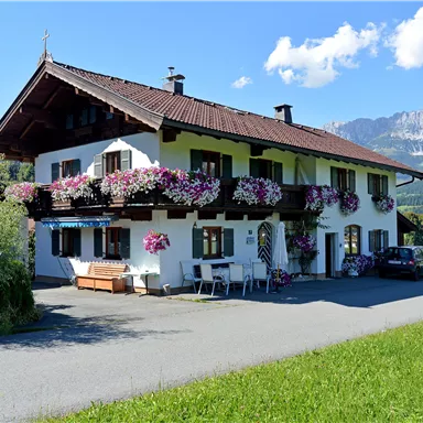 A traditional Bavarian house with blooming flowers on the balconies. The surroundings are green and mountainous, ideal for a summer outing.
