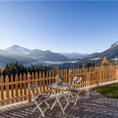 A picturesque view of the mountains and forests. In the foreground, there is a small table and two chairs in front of a pretty fence.