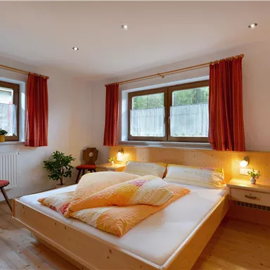 A cozy bedroom with a large bed and colorful pillows. The windows let in plenty of daylight and the orange curtains create a warm atmosphere.