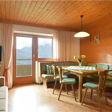 Ein gemütliches Wohnzimmer mit Holzwänden, einem Esstisch und Stühlen. Durch das Fenster sieht man die Berge und den Balkon.
