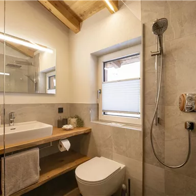 A modern bathroom with a shower, a wooden vanity, and a large mirror surface. The space features bright tiles and a window for natural light.