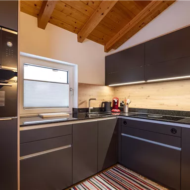 A modern kitchen with wooden ceilings and stylish black furniture. The equipment includes an oven, a sink, and various kitchen appliances.