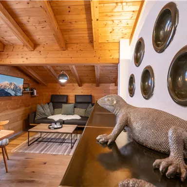 A cozy living room with wooden ceilings, modern furniture, and a TV setup. A decorative lizard rests on a table, and round wall decorations hang on the wall.