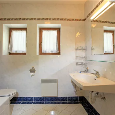 A bright bathroom with a shower, sink, and toilet. Windows with curtains let in light and create a friendly atmosphere.