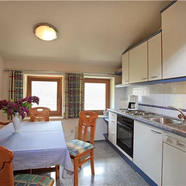 A bright kitchen with a dining table and chairs. A vase with flowers is on the table, and the windows are decorated with curtains.