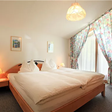 A cozy bedroom with a large double bed and flowering curtains. There is soft light from the table lamps and a view of the outdoor area through the windows.