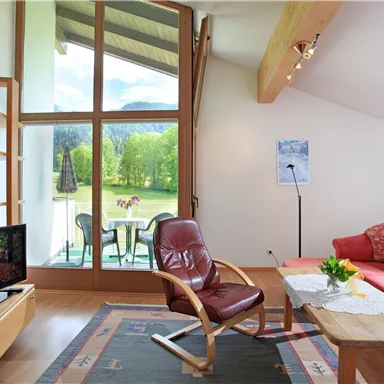 A modern living room with large windows and a view of the green landscape. The furniture is cozy, including a red sofa and a wooden coffee table.