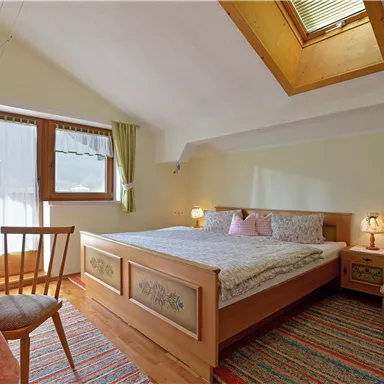 A cozy bedroom with a large bed, a table, and a chair. The room has windows with curtains and a warm, inviting color tone.