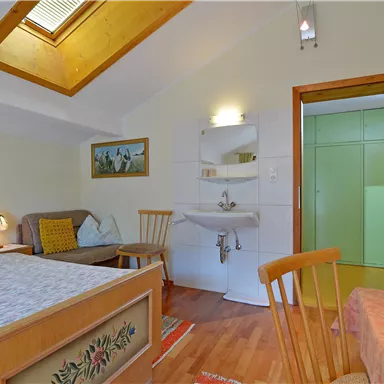 A cozy room with a double bed, chairs, and a washbasin. A bright window provides daylight.
