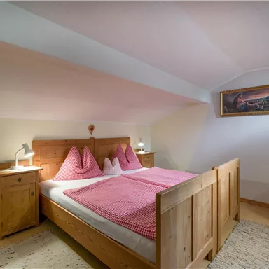 A cozy bedroom with a wooden bed and pink pillows. Next to the bed are nightstands and a chair in a bright room.