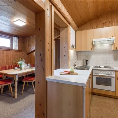 A cozy kitchen with wooden paneling and a dining table. The facilities include a stove and a sink, connected to an adjoining living area.