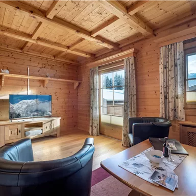 A cozy living space with wooden ceilings and warm colors. In the foreground are two black armchairs and a table with magazines, while a television is visible in the background.