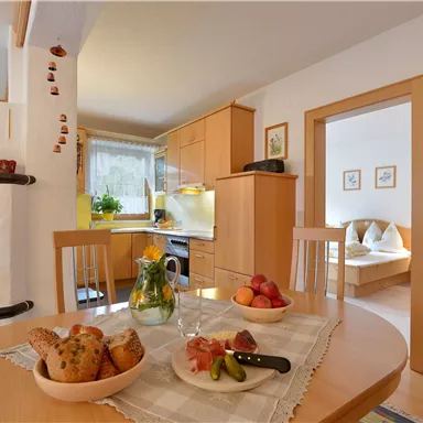 A cozy kitchen with a wooden table and fresh fruit on the table. In the background, kitchen furniture and an adjoining room are visible.