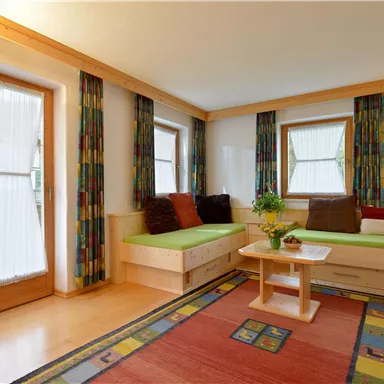 A bright living room with cozy seating and large windows. The room is decorated with colorful curtains and a carpet.