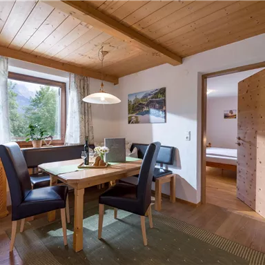 A cozy dining area with a wooden table and black chairs. In the background, windows with a view and another room are visible.