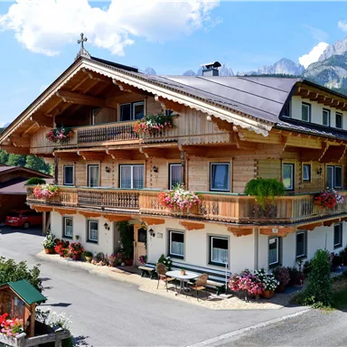 A traditional alpine inn with a wooden construction and colorful flowers on the balconies. In the background, majestic mountains and a blue sky are visible.
