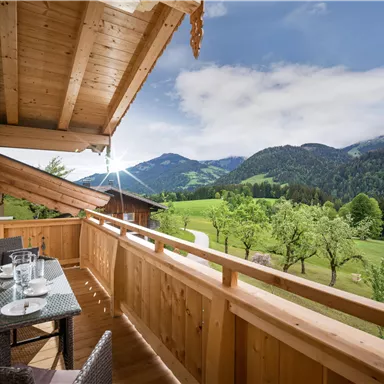 Ein schöner Balkon mit Holzgeländer und einem Tisch für zwei. Im Hintergrund sind grüne Wiesen und Berge unter einem klaren Himmel zu sehen.