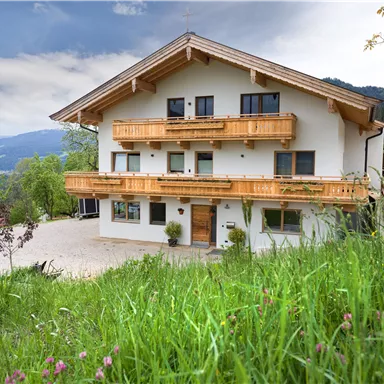 Ein modernes Haus im alpinen Stil mit Holzbalustraden und großzügigen Balkonen. Umgeben von grünen Wiesen und bergiger Landschaft.