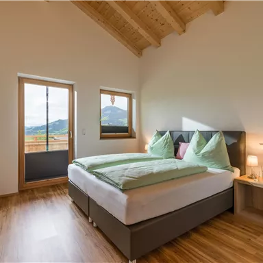 Ein modernes Schlafzimmer mit einem großen Bett und hellen Kissen. Durch die Fenster hat man einen Blick auf eine malerische Landschaft.